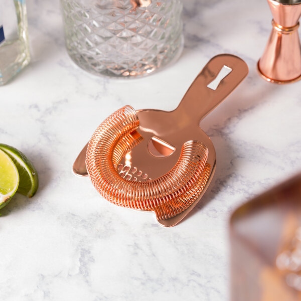 A copper Barfly Hawthorne strainer in a glass on a marble surface.