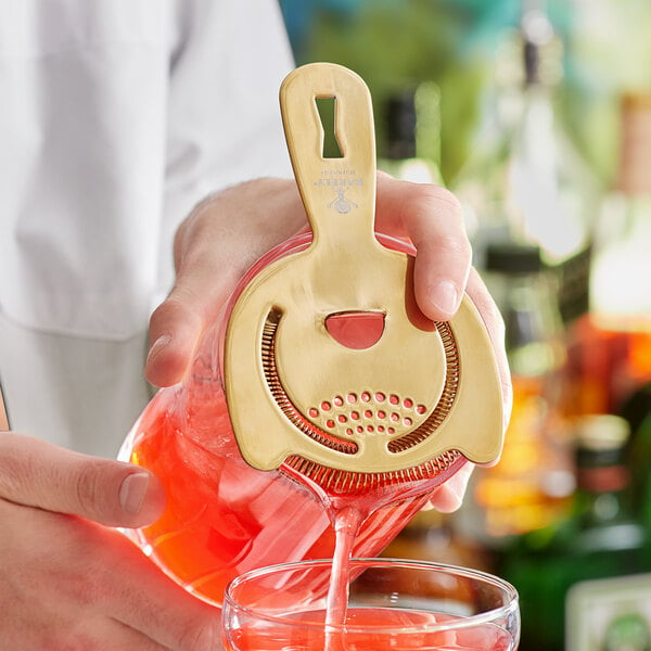 A hand using a Barfly gold-plated Hawthorne strainer to pour a red cocktail into a glass.