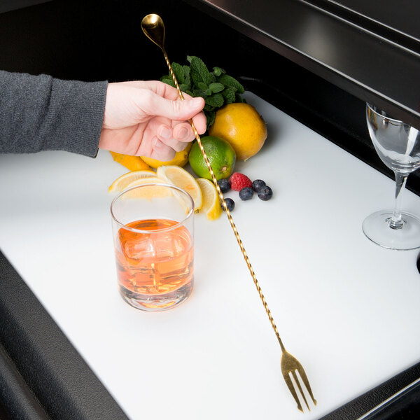 A hand holding a gold Barfly spoon with a fork end next to a glass of orange juice.