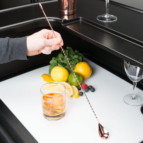 A person using a Barfly copper plated classic bar spoon to stir a cocktail on a tray with fruit and a glass of amber liquid.