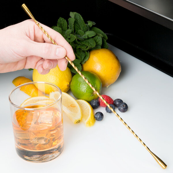 A person using a Barfly gold plated double end stirrer to stir a glass of liquid with ice and lemon slices.