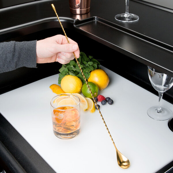 A hand using a Barfly gold plated classic bar spoon to stir a cocktail with ice and a lemon wedge.
