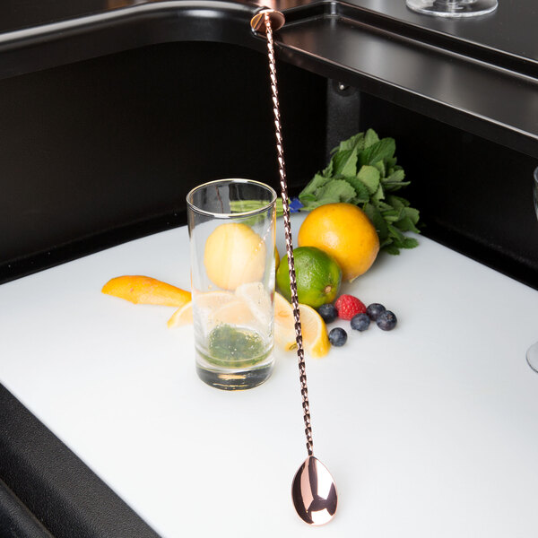 A copper plated Barfly bar spoon with a muddler end stirring a glass of fruit on a white surface.