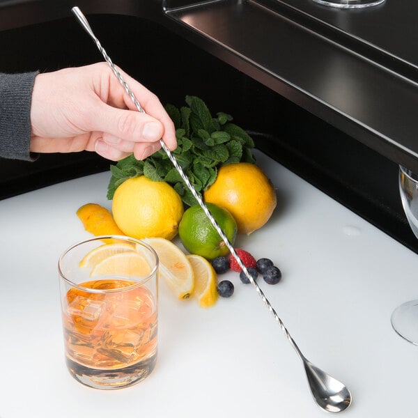 A hand using a Barfly stainless steel bar spoon to stir a cocktail with a lemon wedge.