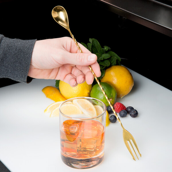 A hand holding a Barfly gold spoon over a glass of liquid with ice and lemon slices.