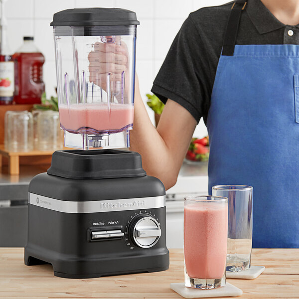 A person in a blue apron using a KitchenAid matte black blender on a wooden surface to make a pink smoothie.