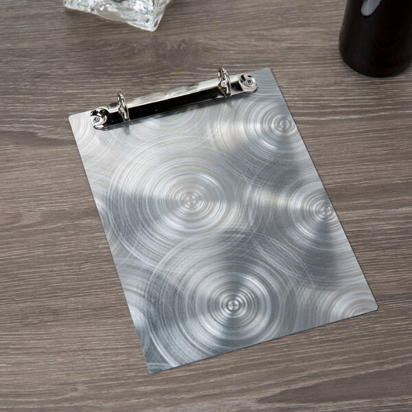 A Menu Solutions Alumitique aluminum menu board with swirl finish and rings on a table.