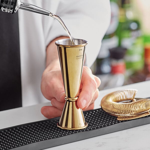 A person using a Barfly gold-plated Japanese style jigger to pour liquid into a gold cup.