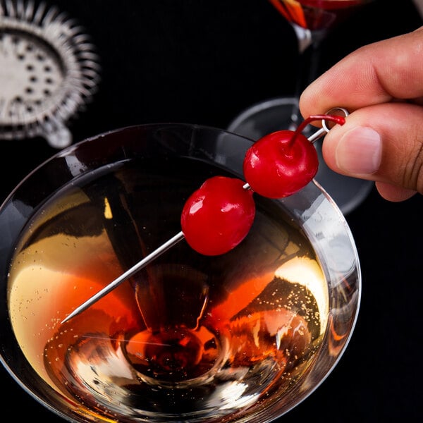 A hand holding a red cherry on a Barfly stainless steel cocktail pick in a drink.