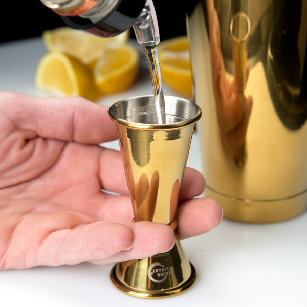 A person using a Barfly gold-plated Japanese style jigger to pour a drink into a small gold cup.