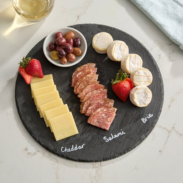 A round black slate tray displaying cheddar cheese, salami, brie cheese, strawberries, and a bowl of mixed olives.