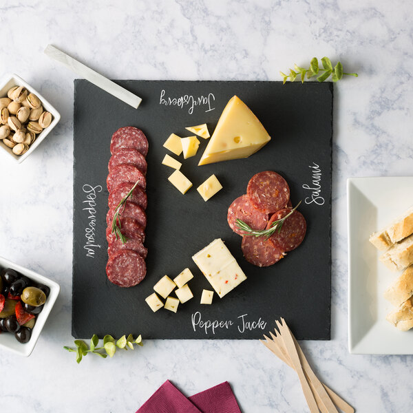 A table with an Acopa black slate tray holding a cheese board, meats, bread, olives, peppers, and pistachios.