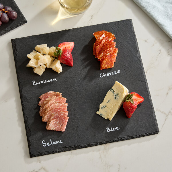 A square black slate tray with four sections labeled for different cheeses and meats, accompanied by soapstone chalk.