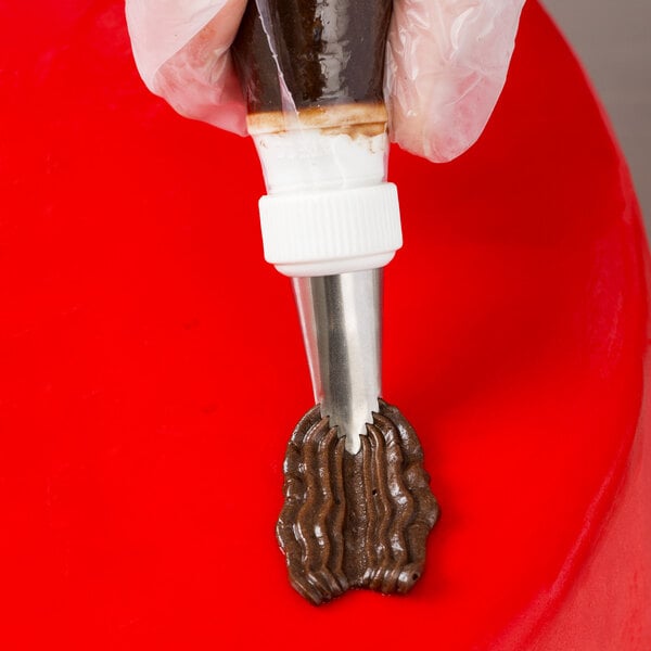 A hand holding an Ateco pastry bag with brown frosting over a red surface.