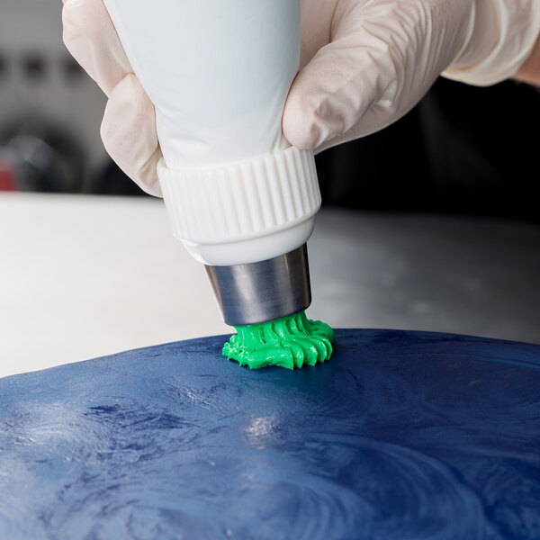 A gloved hand using a white Ateco Russian piping tip with green frosting to frost a blue cake.