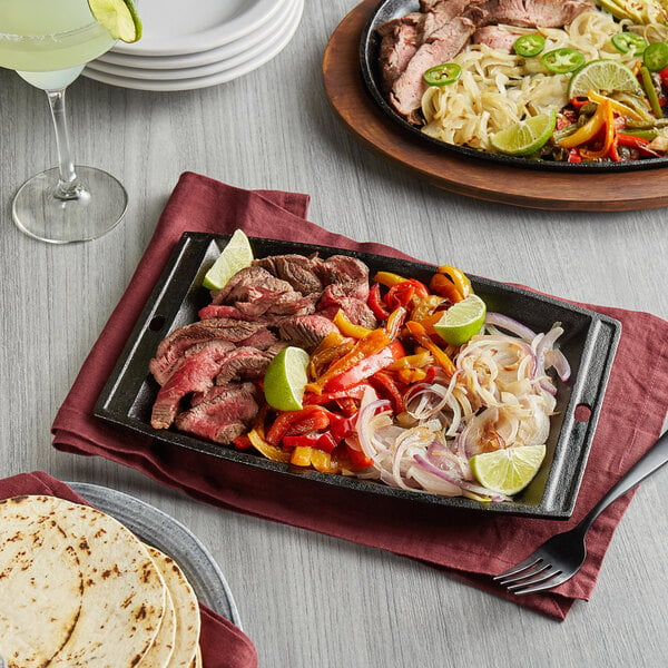 A rectangular cast iron fajita skillet with steak and vegetables on a wood surface.
