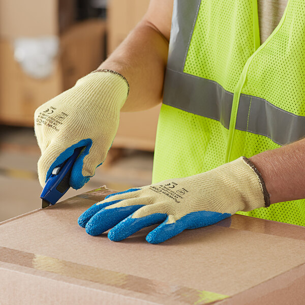 A person wearing Cordova yellow cut resistant gloves with blue latex palm coating and a safety vest using a cutter to cut a box.