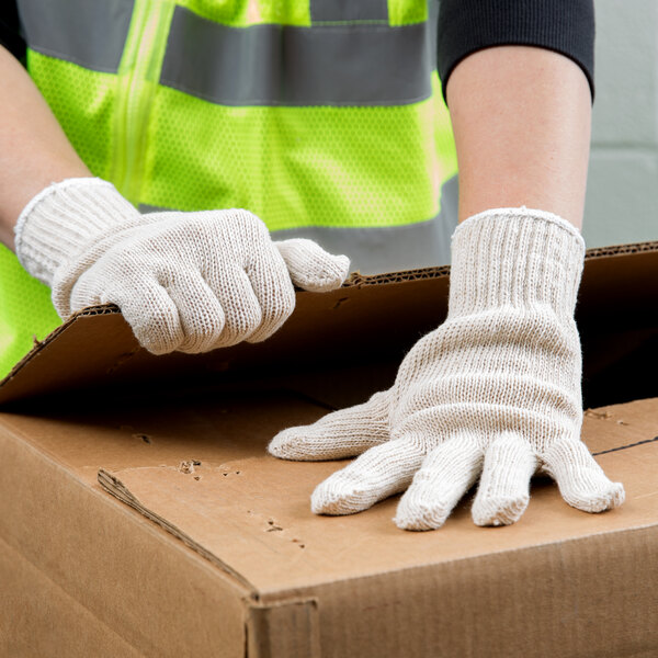 A person wearing Cordova economy weight natural polyester/cotton work gloves and a white shirt is holding a cardboard box.
