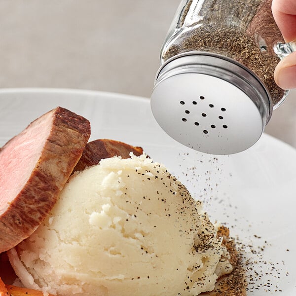 A hand using an Acopa mini mason jar salt shaker to sprinkle salt on a plate of food.