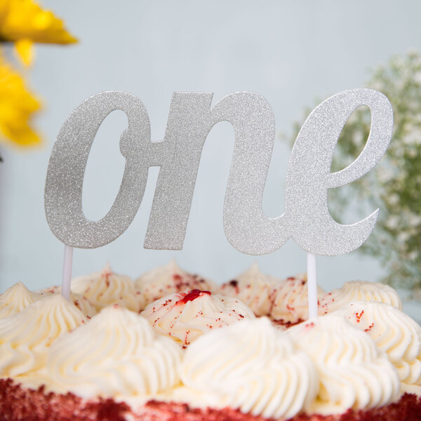A cake with a silver glitter "One" topper on white frosting.