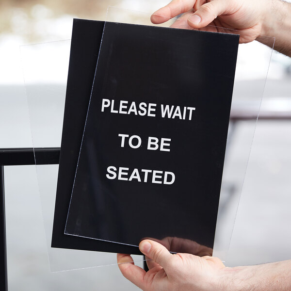A person using a Lancaster Table & Seating stanchion to hold a sign with white text that says "Please wait to be seated"