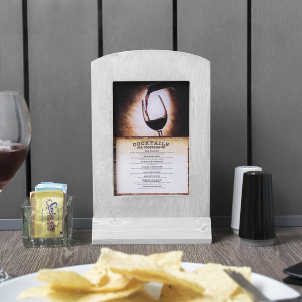 A Menu Solutions white wash wood menu frame on a table with a glass of wine and plate of chips.