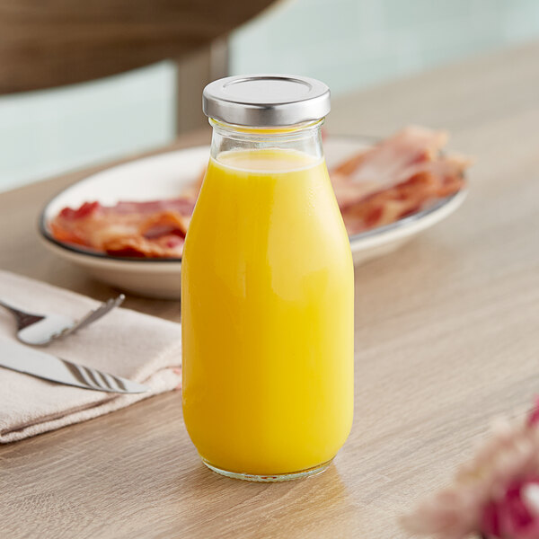 A clear glass milk bottle with a metal lid, filled with orange juice, placed on a table.