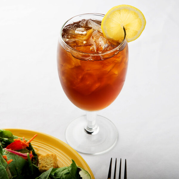 A Libbey Iced Tea Glass filled with brown iced tea on a table with a salad and a lemon slice.