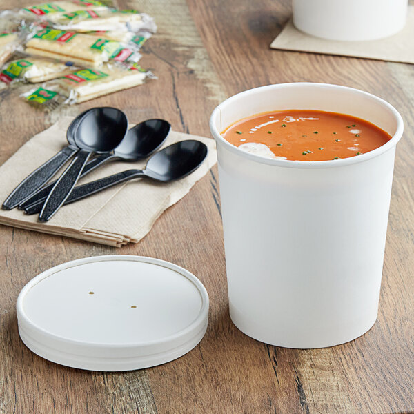 A white Choice paper food container filled with red soup on a table with black spoons.