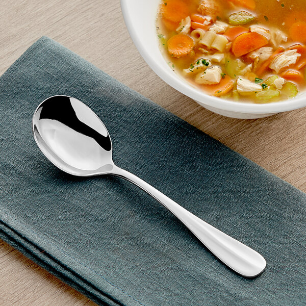An Acopa Benson stainless steel bouillon spoon on a napkin next to a bowl of soup.