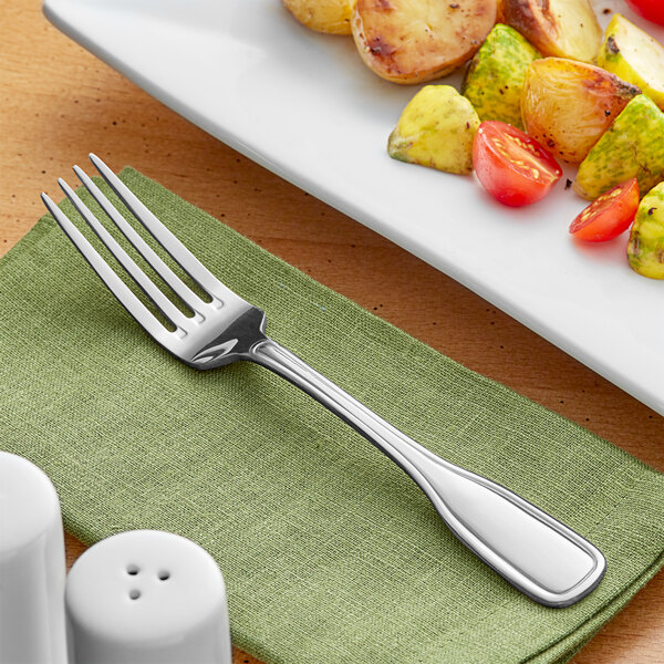An Acopa Saxton stainless steel dinner fork on a napkin next to a plate of vegetables.