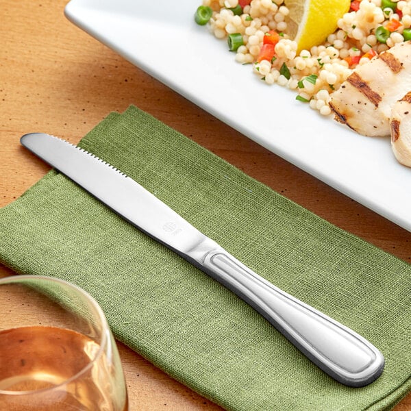 An Acopa Saxton stainless steel dinner knife on a napkin next to a plate of food.