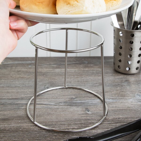 A person using a Choice round metal display stand to hold a plate of bread on a wood table.