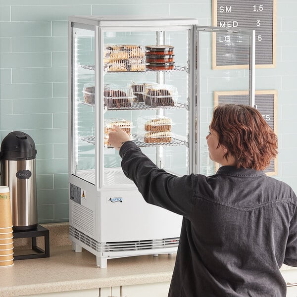 A woman looking at a display of food in a white glass sided countertop display refrigerator.