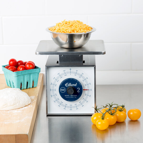An Edlund stainless steel portion scale on a counter with a bowl of cheese in it and tomatoes on a cutting board.