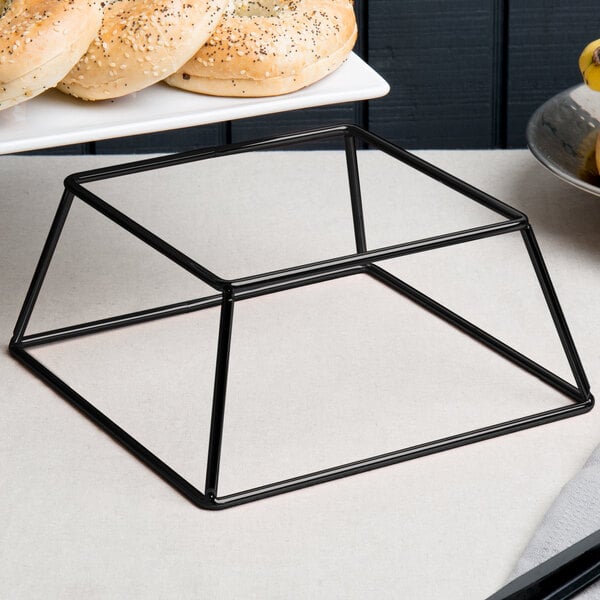 An Acopa black metal square display stand holding a tray of bagels on a white table.
