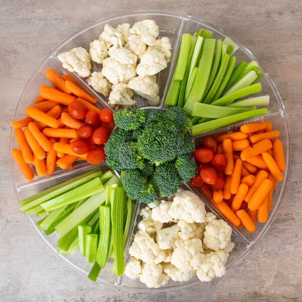 A Fineline clear plastic round tray with 7 compartments of vegetables including carrots, celery, and broccoli.