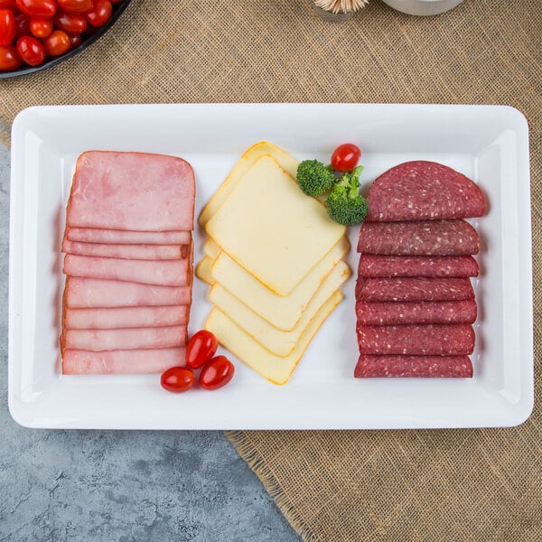 A white plastic rectangular catering tray with a stack of sliced ham, cheese, and a bowl of cherry tomatoes.