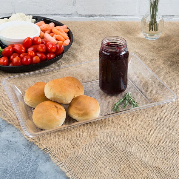 A Fineline clear plastic rectangular catering tray with rolls, bread, and a jar of jam on a table with a bowl of carrots and tomatoes.