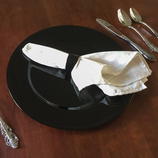 A Tabletop Classics by Walco black plastic charger plate with silverware and a napkin on a table.