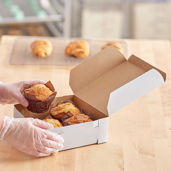 A person in gloves holding a white bakery box filled with muffins.