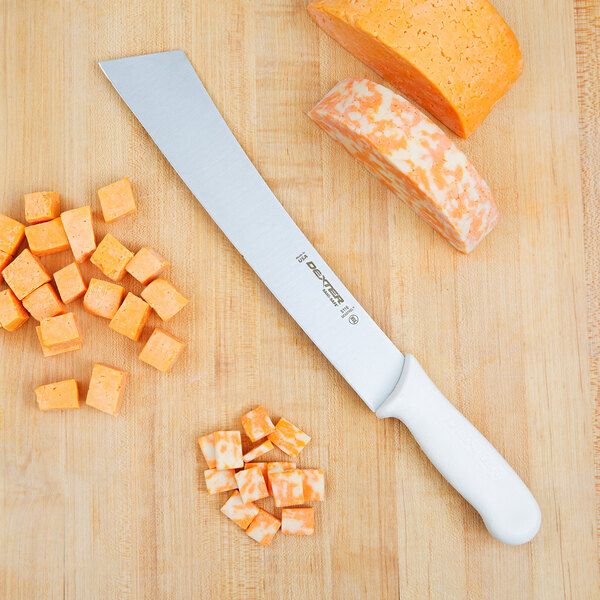 A Dexter-Russell cheese knife next to a block of cheese and cubes of cheese on a wooden cutting board.