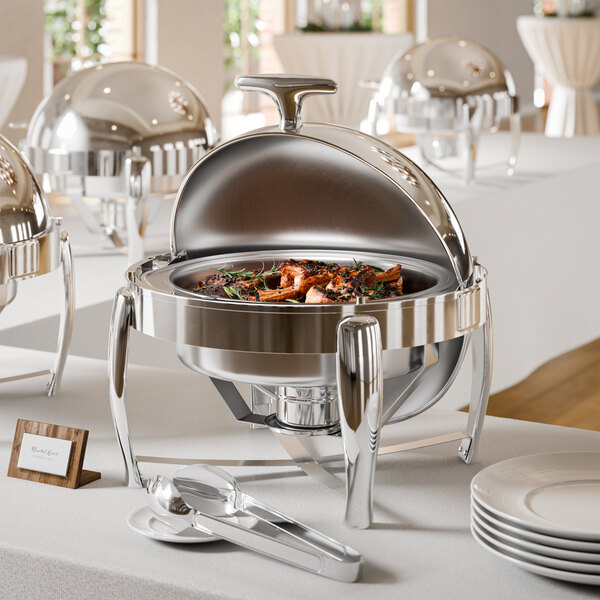 A round stainless steel chafing dish with a roll-top lid is displayed on a white tablecloth, containing cooked food and accompanied by serving utensils and plates.