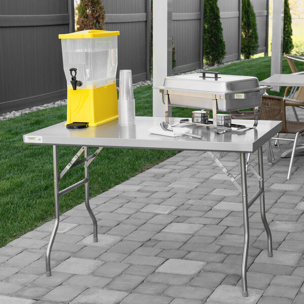 A Regency stainless steel folding work table on a patio with a cooler and cups on it.