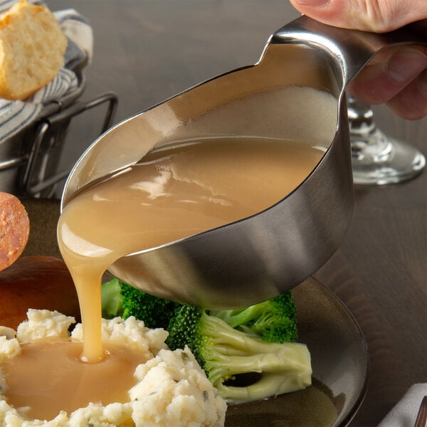 A person using a Tablecraft stainless steel gravy boat to pour gravy over mashed potatoes on a plate.