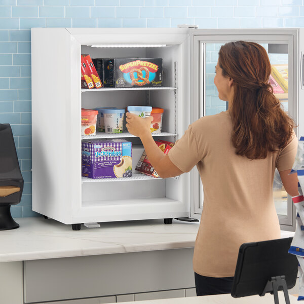 A white countertop display freezer with a swing door, stocked with various frozen foods and being accessed by a person.