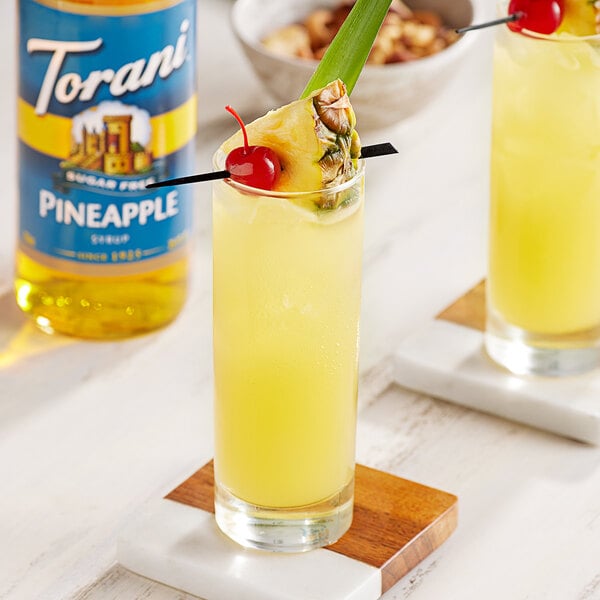 A glass of yellow liquid with a pineapple and cherry garnish.