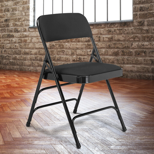A National Public Seating black metal folding chair with a midnight black fabric padded seat in front of a brick wall.