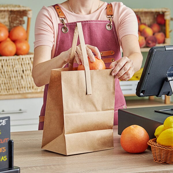 A woman in a pink apron using a Choice brown paper bag to put oranges on a table.