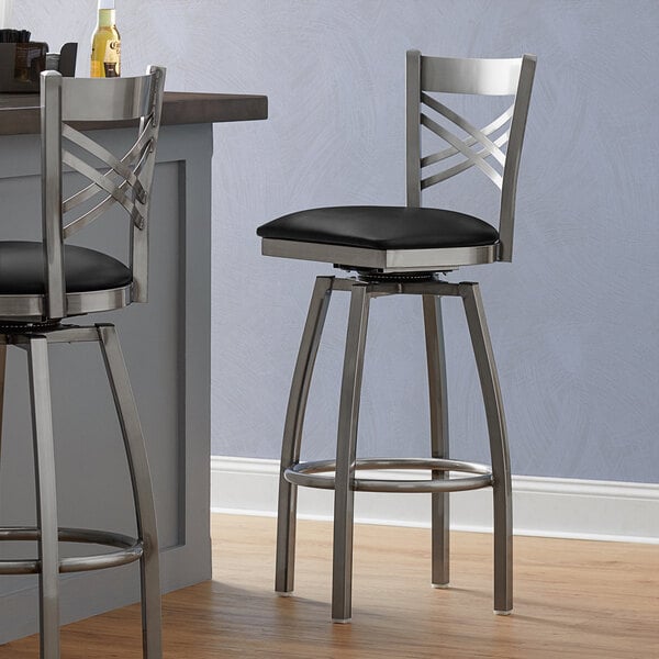 Two Lancaster Table & Seating clear coat finish cross back swivel bar stools with black vinyl padded seats at a counter.